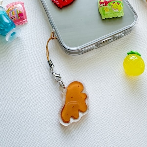 May include: A clear phone case with a brown dinosaur charm attached to it. The charm is on a gold chain with a silver clasp. The phone case has a clear back and a white front. There are other charms on the phone case, including a red heart, a green and yellow package, and a yellow pineapple.