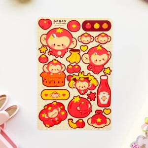 May include: A sticker sheet featuring cartoon monkeys, tomatoes, bananas, and stars. The monkeys are red with yellow accents. The text "BANJO THE TOMATO MONKEY" is at the top. A ketchup bottle is also included.