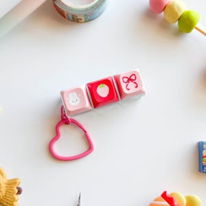 Może przedstawiać: Różowy brelok do klawiatury z trzema klawiszami z ilustracjami królika, truskawki i kokardy. Brelok ma różowy pierścień w kształcie serca. Klawisze znajdują się na białej powierzchni z innymi kolorowymi przedmiotami.