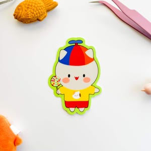 Op de afbeelding: Een kleurrijke sticker met een stripfiguur met een geel shirt, een rode korte broek en een blauw, rood en witte hoed. Het personage houdt een lolly vast. Een geel visvormig object en een roze pincet zijn ook zichtbaar.