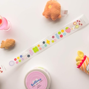 Könnte beinhalten: Eine transparente dekorative Klebebandrolle mit bunten Knöpfen und Fruchtdesigns. Das Klebeband weist eine Vielzahl von Formen und Farben auf, darunter Rosa, Grün, Gelb und Blau. Eine rosa Klebebandrolle mit dem Wort "buttons" ist ebenfalls sichtbar.