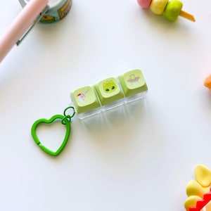 May include: A set of three light green keyboard keycaps with cartoon illustrations of a cat, an alien, and a UFO. The keycaps are held in a clear plastic case and attached to a green heart-shaped keychain.