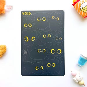 May include: A black sticker sheet with cat-shaped designs, each with yellow eyes. The text "VOID. pako puff" is at the top. A paw print is also included. Small toys surround the sticker sheet.