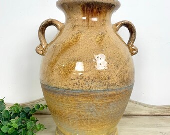 May include: A ceramic vase with a wide mouth and two side handles, featuring a mottled tan and brown glaze on the upper portion and a textured, weathered yellow-brown finish on the lower section, with a distinct horizontal band separating the two.