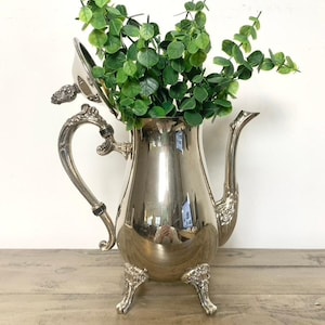 May include: A silver-colored teapot with a curved spout, ornate handle, and three decorative feet. The teapot is filled with green leafy sprigs. The lid is slightly ajar. The teapot is sitting on a wooden surface.
