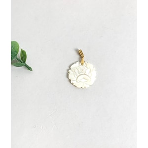 May include: A white, rose-shaped pendant with a gold-colored bail. The pendant is carved with detailed petals and a central rose design. The pendant is set against a white background with a small green plant in the upper left corner.