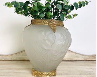 May include: A frosted glass vase featuring a relief floral pattern, accented with an ornate gold-toned metal rim and base. The vase is filled with a green leafy plant, showcasing a clear contrast between the textured glass and metallic trim.