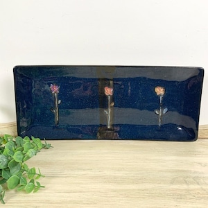 May include: A rectangular, navy blue ceramic dish with three stylized flower designs. The flowers are in shades of pink and orange, with green stems. The dish is on a light wood surface, with greenery in the foreground.