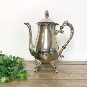May include: A silver-colored coffee pot with a lid and ornate handle. The pot has a curved spout and sits on three decorative feet. The handle is curved and detailed. The lid is topped with a decorative finial. The pot is reflective.
