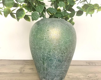 May include: A green vase with a crackle finish holds green leafy branches. The vase has a rounded shape and a textured surface. The background is a light-colored wall and a wooden surface.