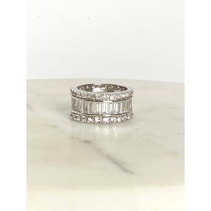 May include: A silver ring with three bands of clear, rectangular-cut stones. The top and bottom bands are lined with smaller, round stones. The ring is set against a white marble surface.