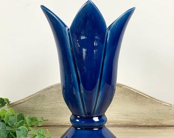 May include: A cobalt blue ceramic vase shaped like a tulip. The vase has a flared base, a rounded midsection, and a tulip-shaped top with pointed petals. The glossy finish reflects light, highlighting the vase's form. The vase is a decorative item.