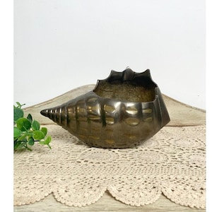 May include: A bronze-colored, seashell-shaped decorative bowl. The bowl has a textured surface and a wide opening. The shell's design includes ridges and a pointed end. It sits on a cream-colored lace doily.