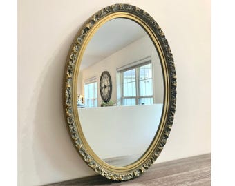 May include: An oval wall mirror featuring a decorative frame with a gold and dark grey finish. The frame has a textured, ornate border with raised detailing. The reflective glass surface shows a room interior with a window and a wall clock.