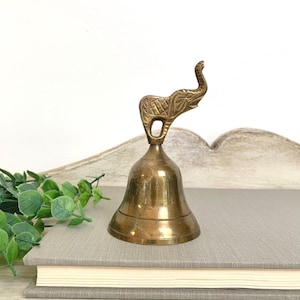 May include: A brass hand bell featuring a decorative elephant figure as the handle. The elephant has etched line details on its body, and the bell has a smooth, polished finish with a flared base. The metal shows a warm, golden-brown tone.