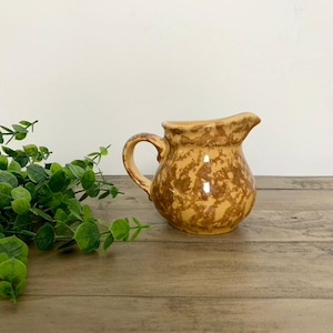 May include: A small, speckled ceramic pitcher with a handle and spout. The pitcher is a warm, earthy brown color with darker brown spots. The handle is curved, and the spout is slightly flared. The pitcher is sitting on a wooden surface.