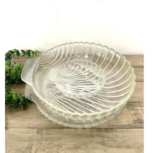 May include: Clear glass serving dish set with a seashell design. The set includes a large, shallow dish and a smaller, round dish. The dishes have a textured surface and a scalloped edge. The set is displayed on a wooden surface.