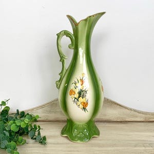 May include: A tall, green and cream-colored ceramic pitcher vase featuring a decorative handle and a floral decal of yellow daffodils on the front. The vase has a flared, scalloped rim with gold-toned trim and a wide, ornate base.