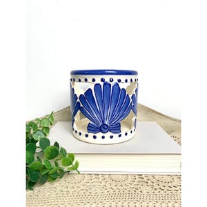 May include: A white ceramic candle holder with a blue seashell design. The holder has cut-out seashell shapes and a blue rim. The candle holder is sitting on a stack of books.