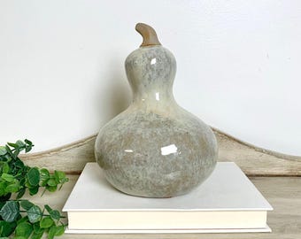 May include: A ceramic gourd-shaped sculpture with a glossy, mottled finish in shades of cream, beige, and soft gray. The piece features a smooth, rounded base that tapers into a narrow neck, topped with a small, curved, unglazed tan stem.