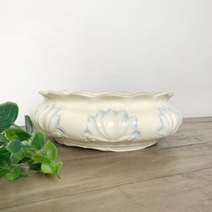 May include: Cream-colored ceramic bowl with a scalloped rim and blue floral designs. The bowl has a wide, shallow shape, suitable for decorative or functional use. The floral pattern is repeated around the exterior of the bowl.