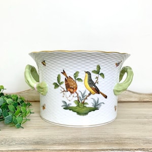 May include: A white porcelain cachepot with a textured basket-weave pattern, featuring two light green scrolled handles and a gold-trimmed rim. The exterior is decorated with a colorful illustration of two birds perched on a branch and butterflies.