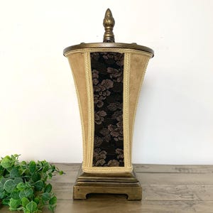 May include: A decorative, vintage-style urn with a square base. The urn features a tan fabric exterior with a central panel of black floral lace. Gold trim and a decorative finial top the urn, which is likely used for home decor.
