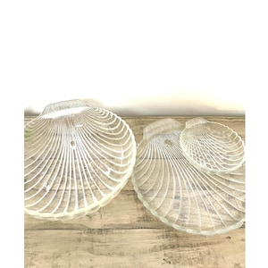 May include: Clear glass seashell-shaped serving dishes. The dishes have a ridged design and are arranged on a light-colored wooden surface. The set includes three different sizes, perfect for serving appetizers or displaying decorative items.