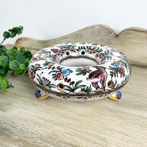 May include: A ceramic, ring-shaped decorative piece with a white base, adorned with colorful floral and bird motifs. The piece rests on three small, rounded feet and features a detailed, hand-painted design. The overall aesthetic is vintage and artisanal.