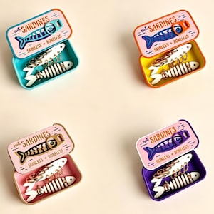 May include: Four rectangular metal tins with lids featuring a sardine graphic and the text not SARDINES SKINLESS + BONELESS. Each tin contains a white fish skeleton shaped jewelry holder with gold-toned hoop earrings and chain links inside.