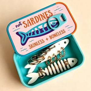 Könnte beinhalten: Eine rechteckige türkisfarbene Dose mit einem rosafarbenen Deckel, der eine blaue Fischgrafik und den Text not SARDINES SKINLESS + BONELESS zeigt. Darin befinden sich zwei weiße, fischförmige Schmuckhalter aus Holz mit goldenen Creolen.