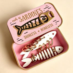 以下が含まれることがあります： 「not SARDINES」「SHINLESS + BONELESS」と書かれたプルタブ付きのピンク色の金属缶。中には魚の形をした白いジュエリーディスプレイが入っており、ゴールドカラーのフープピアスと魚の骨のチャームが付いたゴールドカラーのチェーンネックレスが飾られています。