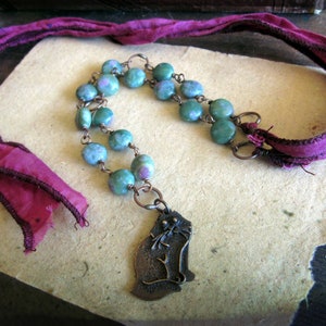 May include: A green and pink gemstone necklace with a copper chain and a copper cat pendant. The necklace is on a burgundy silk ribbon.