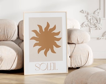 May include: Framed art print featuring a stylized sun design in shades of brown and beige. The word "SOLEIL" is printed in a matching brown font below the sun illustration. The artwork is displayed in a light-colored wooden frame.