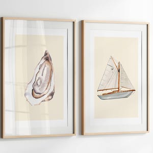 以下が含まれることがあります： 2つの額入り水彩画プリント。1つは、茶色と白の色合いの牡蠣の殻が特徴です。もう1つは、白い帆、灰色の船体、木製のアクセントを備えたヨットを描いています。どちらのプリントも明るいベージュの背景で、明るい木製のフレームに入っています。