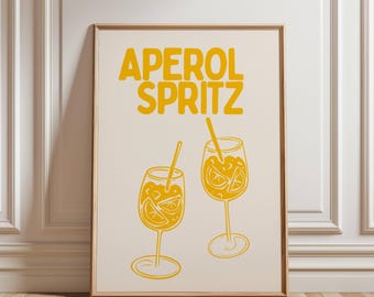 May include: A framed print featuring the words "APEROL SPRITZ" in yellow block letters above two yellow line-drawn cocktail glasses filled with orange slices and straws. The print has a cream-colored background.