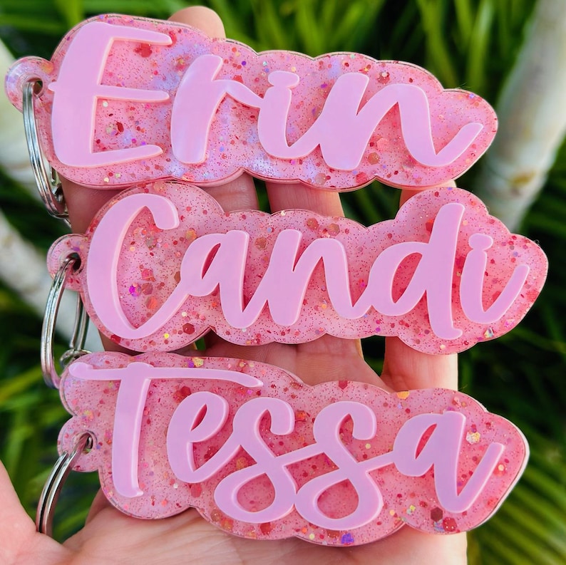 May include: Three custom keychains featuring pink glitter resin bases with scalloped edges. Each keychain displays a name in raised, solid light pink script lettering: Erin, Candi, and Tessa. Each piece includes a silver metal split ring attachment.
