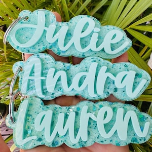 May include: Three custom keychains featuring the names Julie, Andra, and Lauren in raised, light teal script lettering. Each keychain has a cloud-shaped base made of translucent, glittery teal resin and includes a silver metal split ring.