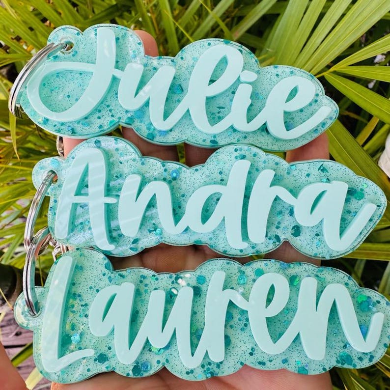 May include: Three custom keychains featuring the names Julie, Andra, and Lauren in raised, light teal script lettering. Each keychain has a cloud-shaped base made of translucent, glittery teal resin and includes a silver metal split ring.