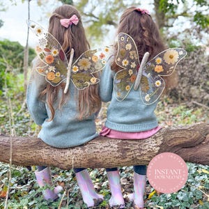 May include: Two children wearing fairy wings decorated with dried flowers, sitting on a log. The wings are clear with a white outline and a wooden centre. They are wearing blue jumpers, pink skirts, and iridescent wellington boots. The image has a soft, natural aesthetic.