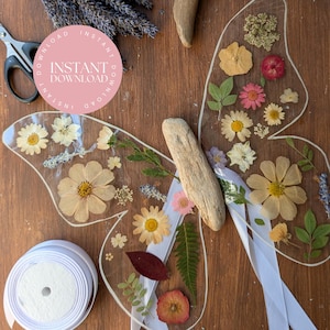 May include: Clear acrylic butterfly wings decorated with pressed flowers and greenery. The wings feature daisies, pink and yellow flowers, and various leaves. White ribbon and a wooden piece are attached. The image includes the text "INSTANT DOWNLOAD".