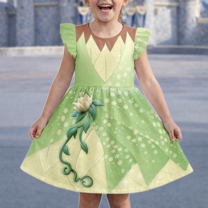 May include: A green and yellow dress with a floral design. The dress features a brown neckline and ruffled sleeves. A large white flower with green vines adorns the front of the dress. The dress is designed for a child.