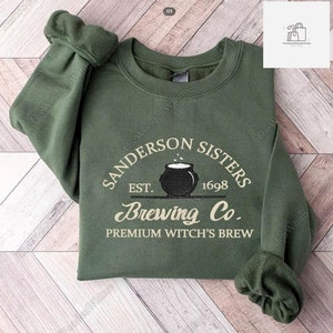 Op de afbeelding: Groen sweatshirt met een wit grafisch ontwerp met de tekst "Sanderson Sisters Est. 1698 Brewing Co. Premium Witch's Brew" met een zwarte ketel en stoom die eruit komt.