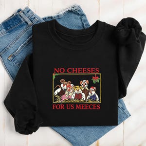 May include: Black sweatshirt with the text "NO CHEESES FOR US MEEECES" in red. The graphic features cartoon mice in festive attire. The sweatshirt is folded on top of a pair of blue jeans.