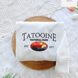 Peut inclure: Sweat-shirt blanc avec un imprimé graphique d'un paysage de coucher de soleil et le texte "TATOOINE NATIONAL PARK EST 1977".