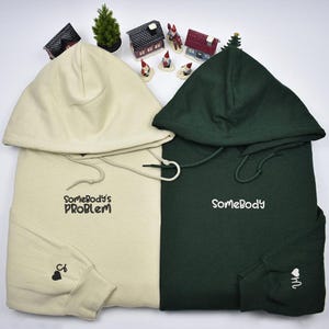 Könnte beinhalten: Zwei Kapuzenpullover, einer cremefarben und einer dunkelgrün, mit gesticktem Text. Der cremefarbene Pullover sagt "Somebody's Problem" und der grüne Pullover sagt "Somebody". Beide Pullover haben ein kleines schwarzes Herz auf dem Ärmel.