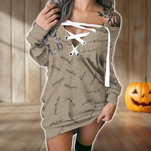 Può includere: Abito beige a maniche lunghe con scollo a V e dettaglio stringato bianco. L'abito è decorato con motivi a punto croce neri. Una zucca arancione intagliata è sullo sfondo, suggerendo un tema di Halloween.