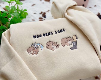 May include: A beige sweatshirt with the text "Moo Deng Gang" embroidered on the front. The sweatshirt features three embroidered cartoon hippos, one with a water spout, one with a closed mouth, and one with a boot on its head.