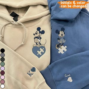 Op de afbeelding: Twee geborduurde hoodies, één beige en één blauw, met Mickey en Minnie Mouse-ontwerpen. Mickey staat op een blauw hartvormig puzzelstuk en Minnie op een wit puzzelstuk. Initialen en kleuren kunnen worden gewijzigd. De beige hoodie heeft de initiaal "K" op de mouw geborduurd.