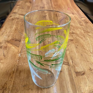 May include: A clear glass drinking glass with a yellow and green floral pattern. The pattern features stylized flowers and leaves.